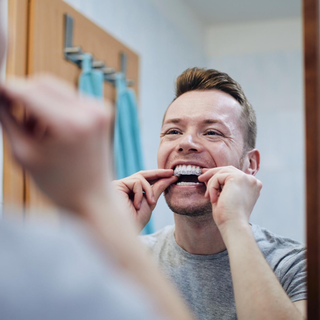 Man smiling and examining clear aligner orthodontic treatment in bathroom mirror.